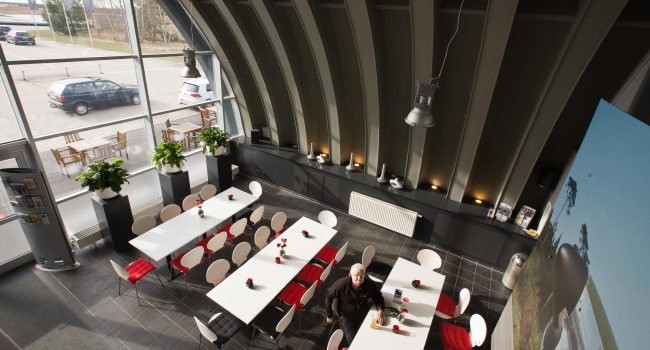 Blick von oben auf ein modernes, lichtdurchflutetes Bistro mit weißen Tischen, roten Sitzkissen und großen Pflanzen vor Panoramafenstern.